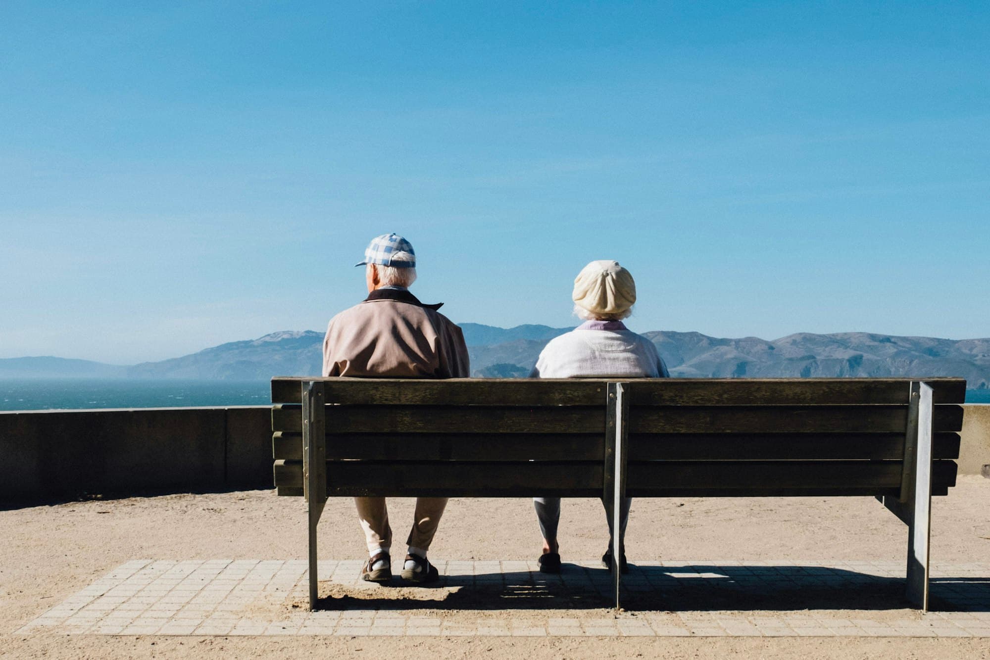 An older couple sitting together on a bench, seen from behind, looking out over the ocean.